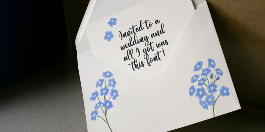 A white envelope with blue flowers containing a white notecard with two blue flowers and the text: "Invited to a wedding and all I got was this font!" The font used for the text is a stereotypical wedding font.